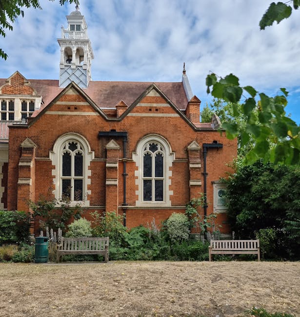 A landscaped front garden in front of a historic red brick church building with arched white-framed windows and decorative stone detailing. The garden features a variety of lush green plants, includin